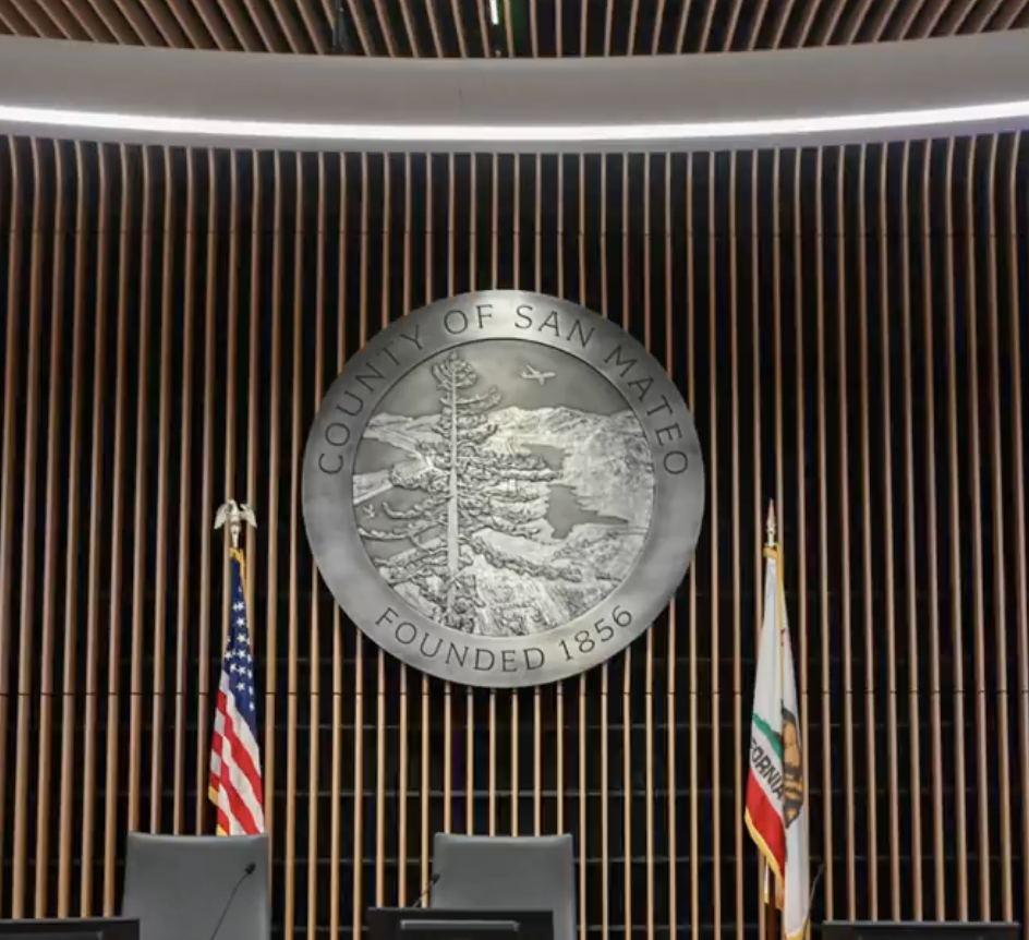 San Mateo County seal displayed behind the Board of Supervisors dais with U.S. and California flags.