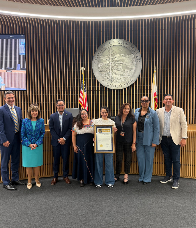 Eight people stand beneath the San Mateo County seal, with a person at the center holding a framed proclamation.