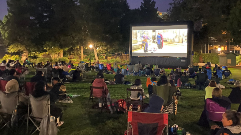 A crowd sits on blankets and lawn chairs in a park at dusk, watching an animated film on a large inflatable outdoor screen.