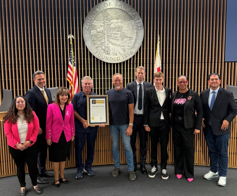 Nine people pose beneath the San Mateo County seal, with one holding a framed proclamation.