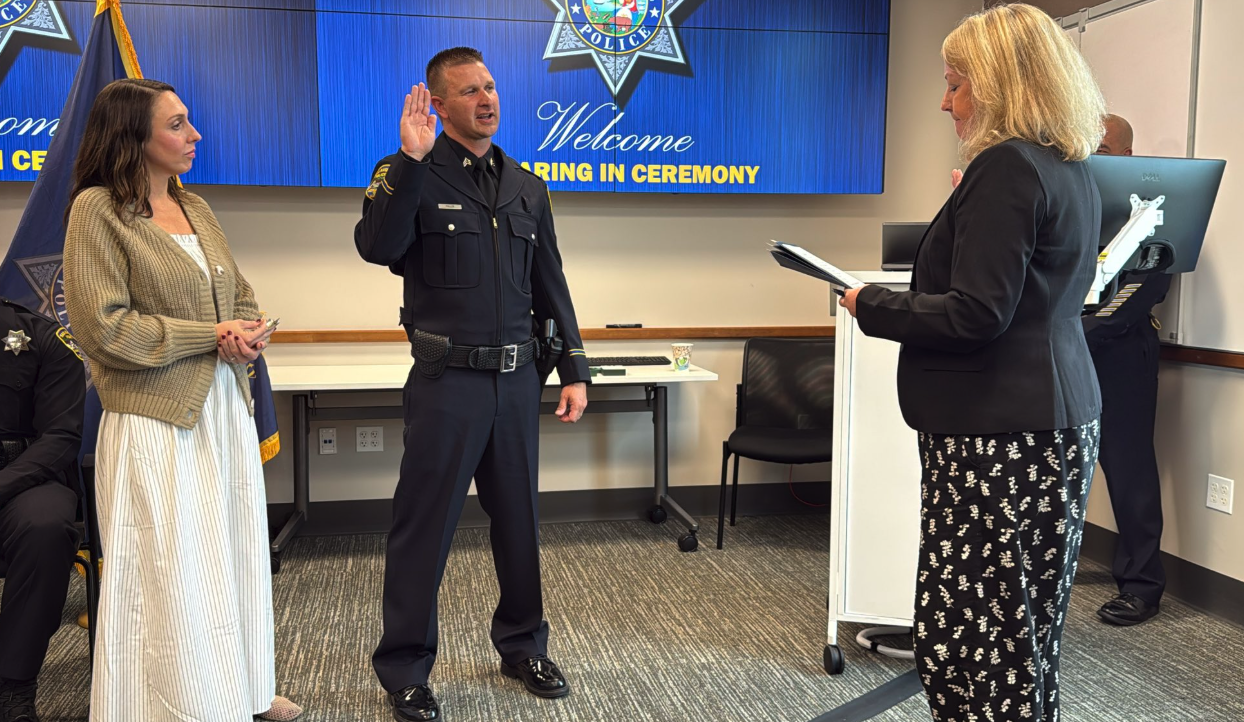 A San Rafael police officer raises his right hand while taking the oath of office during a swearing-in ceremony, as a woman reads from a document and another woman stands beside him.