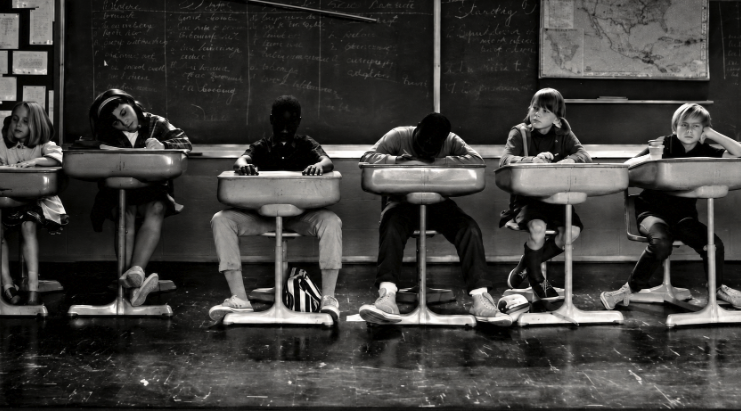 Black-and-white photo of six children with heads down at old-style school desks in a classroom with a chalkboard and map.