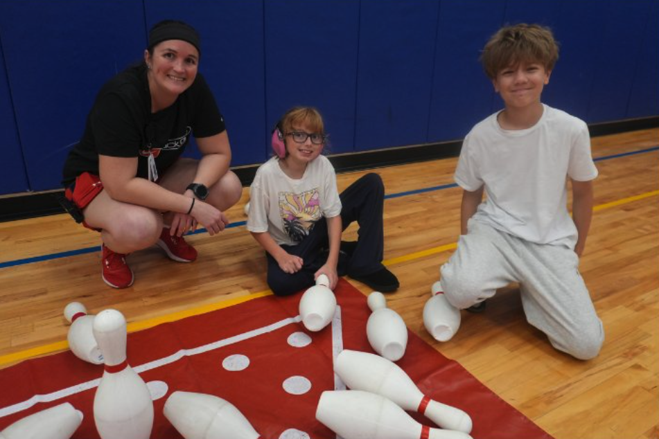 An adult wearing a black staff shirt kneels on a gym floor next to two children, all three smiling, beside a set of oversized foam bowling pins arranged on a red mat for an adaptive bowling activity.