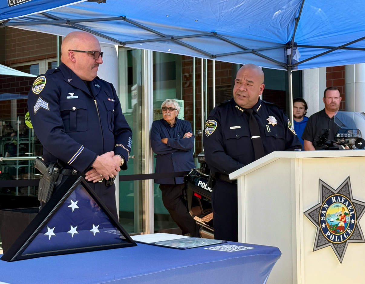San Rafael Police Chief Dave Spiller speaks at a podium bearing the department's badge, standing beside Sgt. Michael Mathis in dress uniform, with a folded American flag displayed on the table during a retirement ceremony.