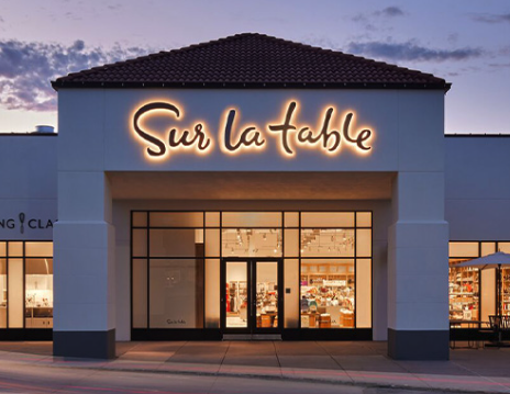 Exterior of a Sur La Table storefront with illuminated sign at dusk.