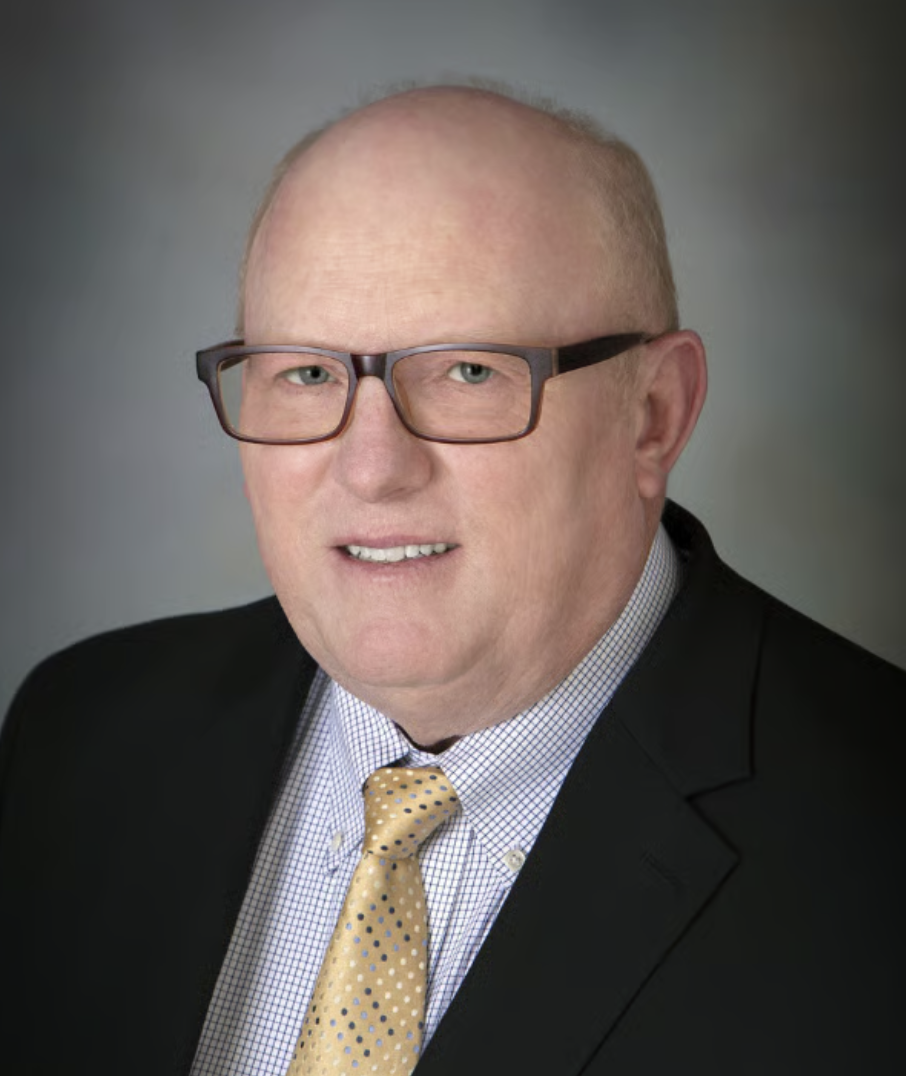 Terence Collins wears glasses, a dark jacket, a checked shirt and a yellow patterned tie against a gray background.
