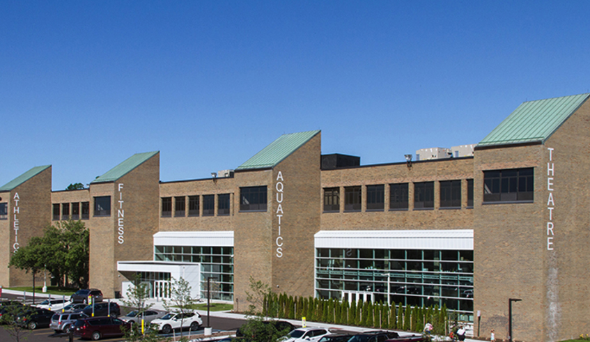 Exterior of The Hawk community center in Farmington Hills, a brick building with sections labeled Athletic, Fitness, Aquatics and Theatre under a blue sky.