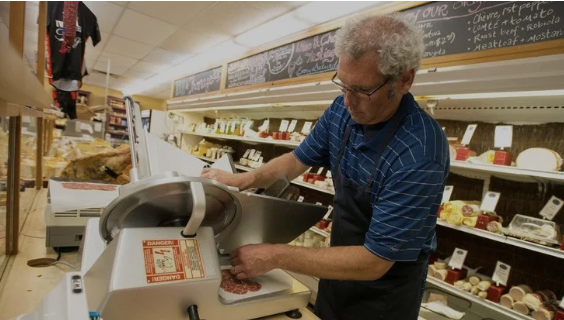 Tommy York slices meat at the deli counter of York Food + Drink, surrounded by shelves of cheese and specialty foods.