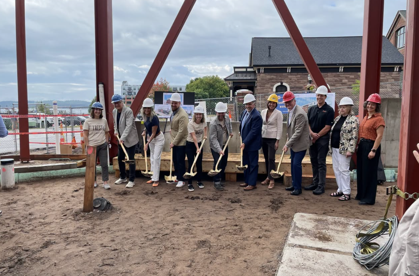 Community members and officials in hard hats hold ceremonial gold shovels at the Trestle Hotel groundbreaking site on Lakeshore Boulevard in downtown Marquette, with steel beams and Lake Superior visible in the background.