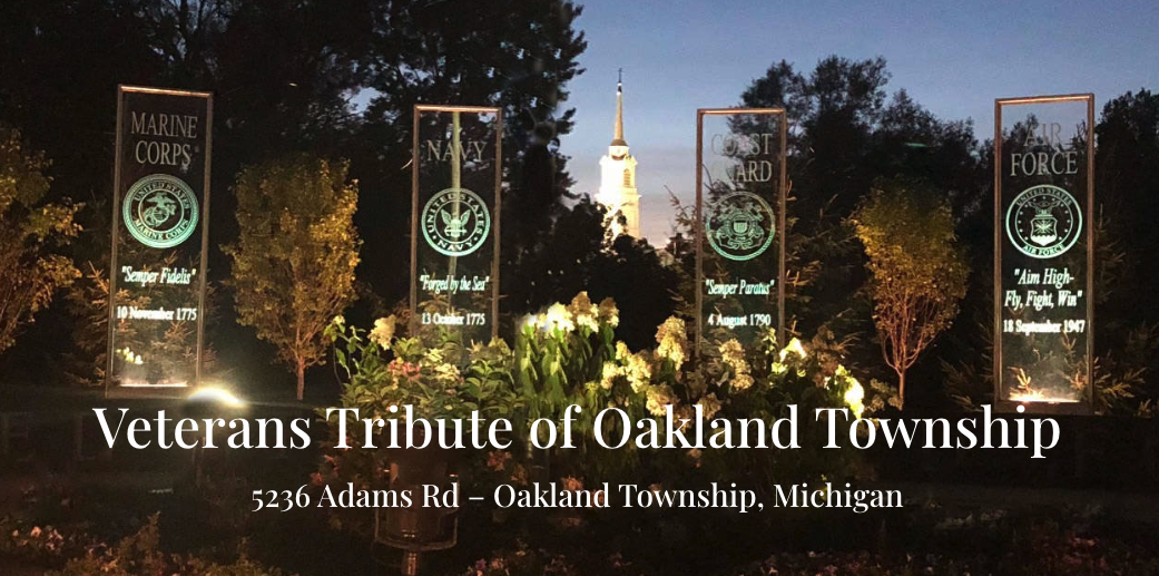 Illuminated memorial banners for the Marine Corps, Navy, Coast Guard and Air Force at the Veterans Tribute of Oakland Township, with a church steeple visible at dusk.