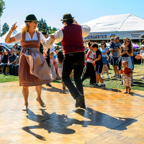 Traditional Bavarian folk dancers perform on the dance floor at Walnut Creek Oktoberfest, with the Biergarten tent visible behind them.
