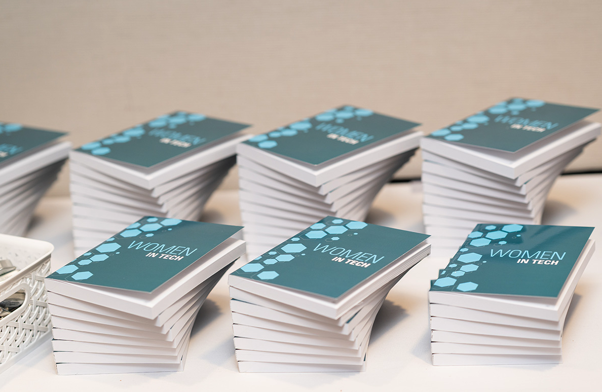 Stacks of teal "Women in Tech" companion workbooks displayed on a table.
